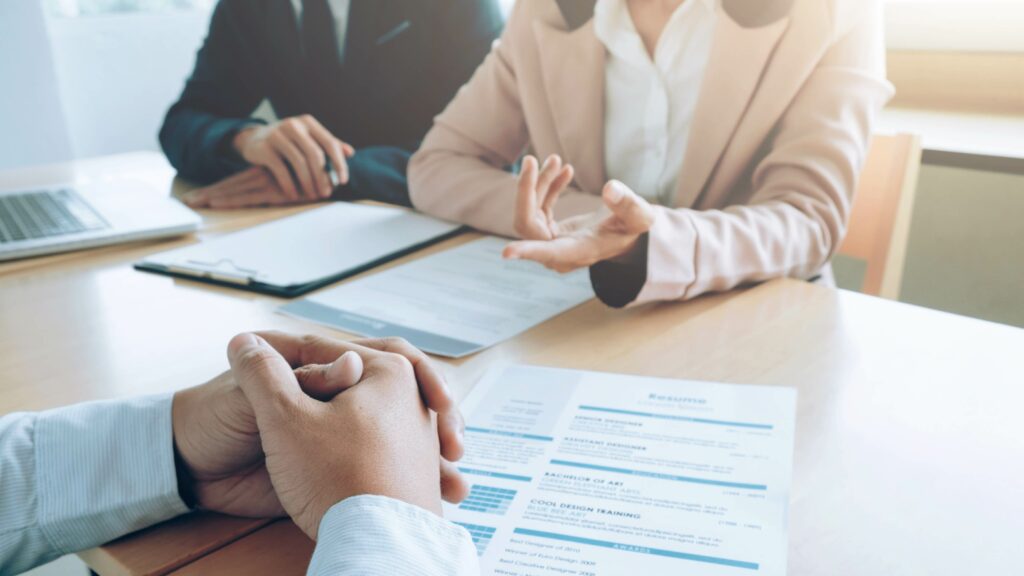 L'image montre une scène professionnelle où deux individus, probablement des recruteurs ou des responsables RH, discutent avec un candidat potentiel lors d'une entrevue d'embauche. Le candidat, assis en face des deux autres personnes, est partiellement visible avec une vue en gros plan sur ses mains jointes. Sur la table, on peut apercevoir des documents, vraisemblablement un CV. L'atmosphère semble professionnelle et sérieuse, avec un décor de bureau moderne.