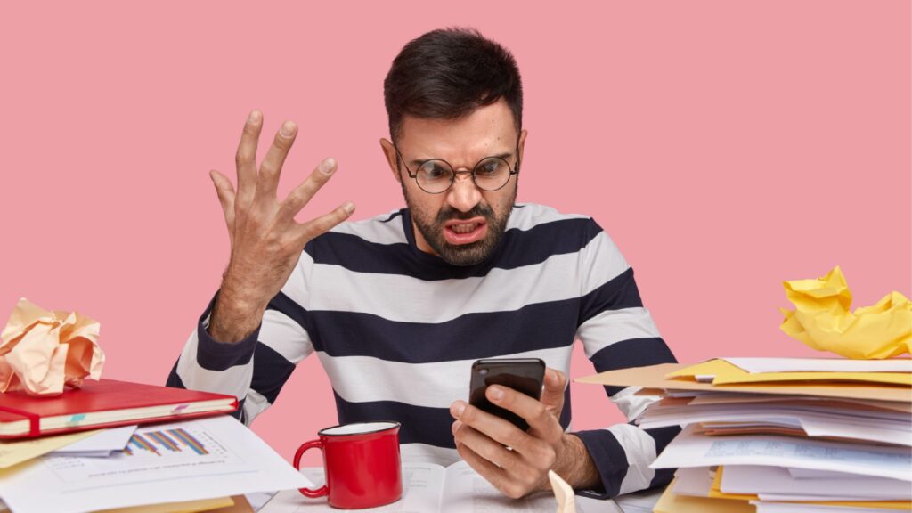 Une personne frustrée assise à un bureau, entourée de papiers froissés, de dossiers ouverts, et d'une tasse rouge, regarde son smartphone avec une expression agacée et gestes des mains montrant de la confusion, sur fond rose.