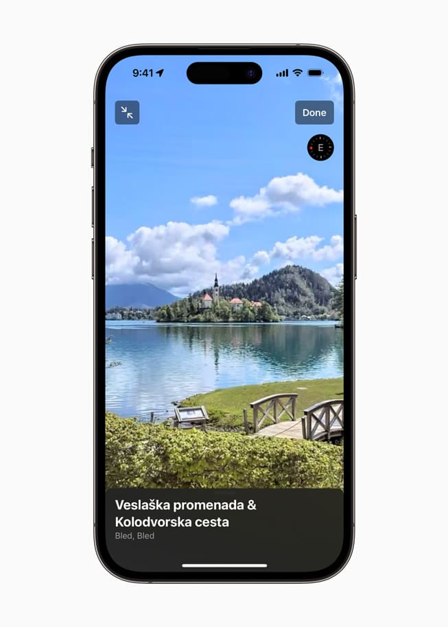 Image montrant l'écran d'un iPhone avec la fonction Look Around d'Apple Maps, offrant une vue interactive au niveau du lac Bled, en Slovénie. On y voit une promenade pittoresque avec des bancs, un lac paisible et une église sur une colline en arrière-plan sous un ciel bleu avec des nuages blancs.