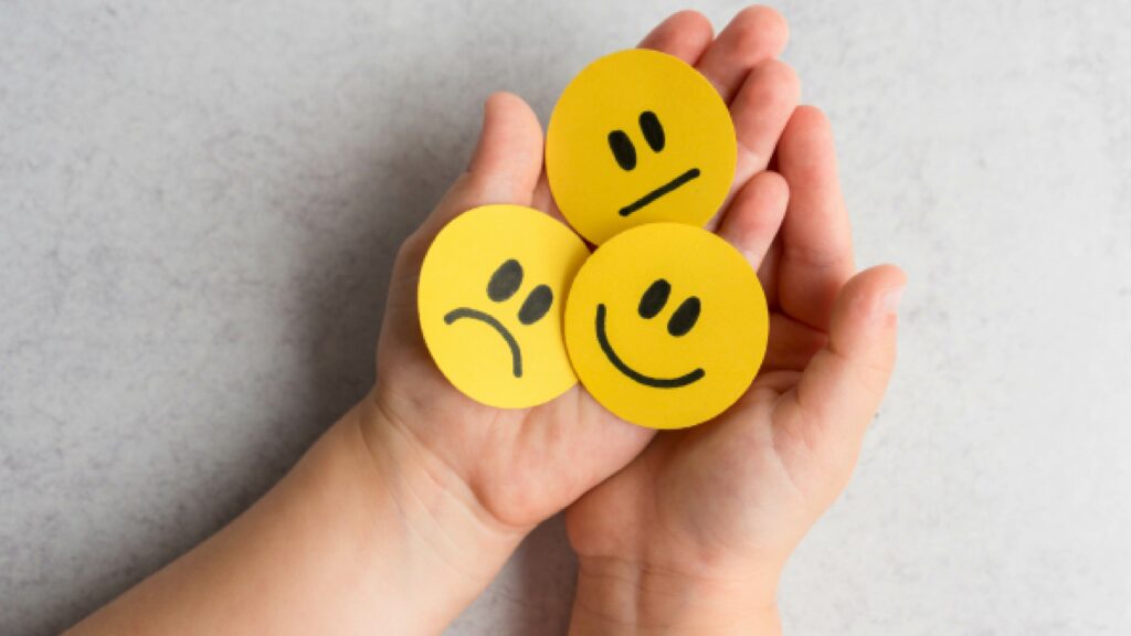 Une personne tenant dans ses mains trois smileys représentant chacun une émotion négative, positive et neutre.
