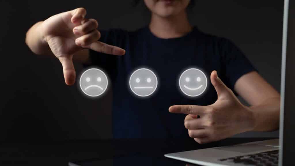 Un homme utilisant un ordinateur portable et une tablette, illustrant la gestion des avis en ligne. Il fait des gestes avec ses mains pour symboliser différentes expressions faciales : une tristesse, une indifférence et une satisfaction, représentées par des icônes de visages.