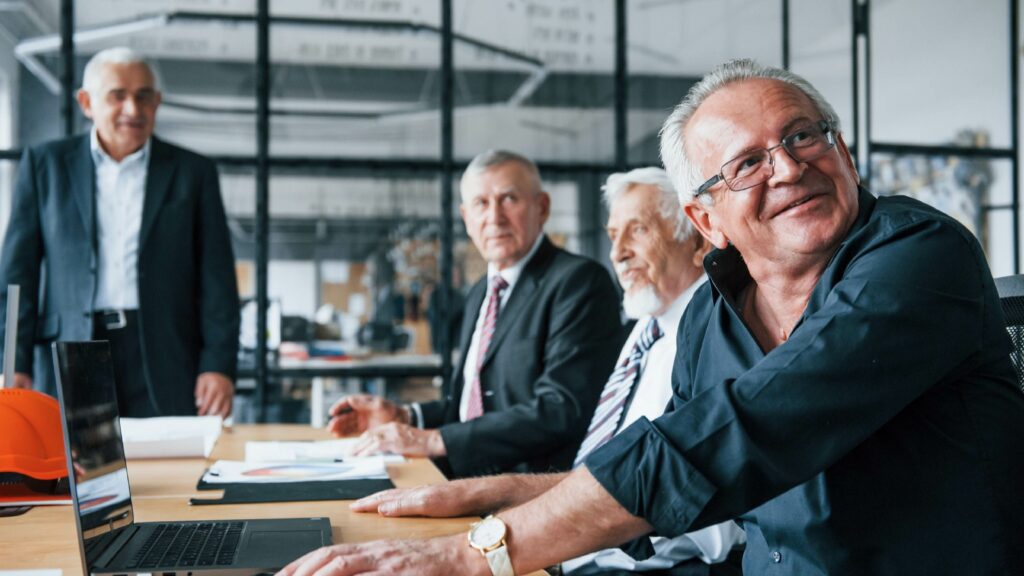 Un groupe de seniors professionnels souriants, collaborant autour d'une table de réunion avec des ordinateurs portables, dans un bureau moderne.