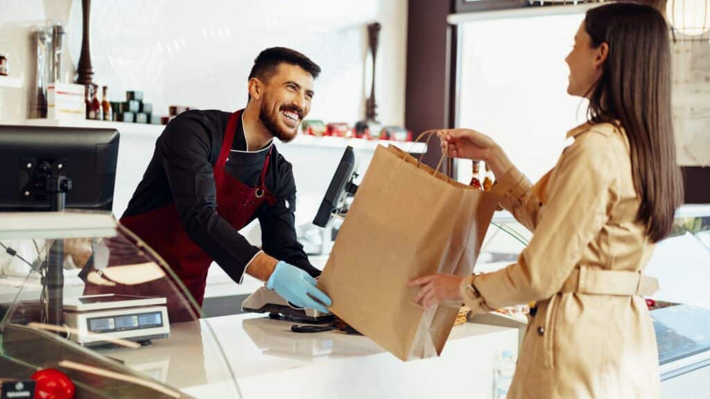 Un vendeur souriant et portant un tablier rouge tend un sac en papier brun à une cliente dans une boutique moderne. La cliente, vêtue d'un manteau beige, accepte le sac avec un sourire, illustrant une expérience client positive et accueillante entre le personnel et les clients.