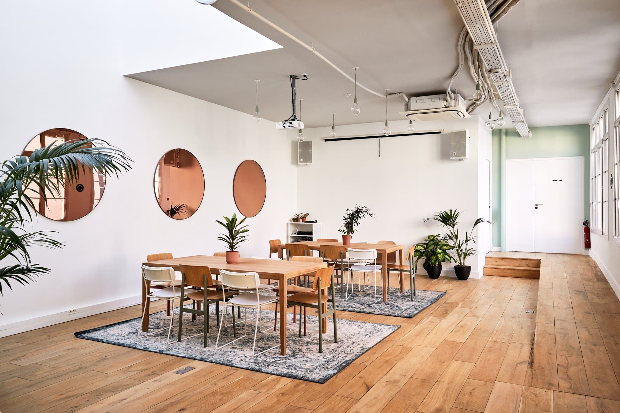 Salle de réunion lumineuse et moderne avec des tables en bois clair, des chaises assorties, et des plantes d'intérieur. Les murs blancs sont décorés de grands miroirs circulaires, ajoutant une touche design à l'espace. Le sol en bois et les tapis doux complètent l'ambiance chaleureuse et accueillante.