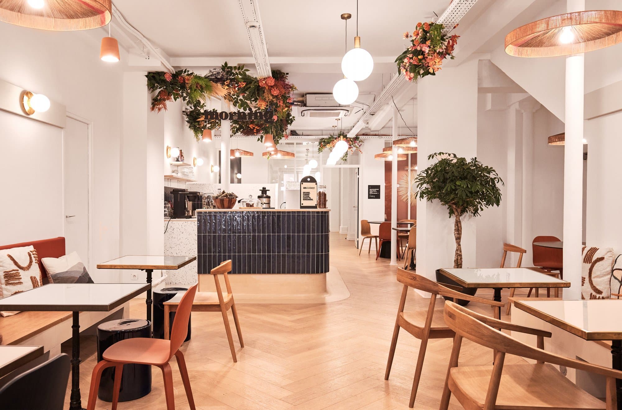 Espace de restauration élégant avec un comptoir central bleu marine, décoré de plantes suspendues et de fleurs. L'éclairage est chaleureux avec des suspensions modernes, et l'agencement des tables et des chaises en bois clair crée une atmosphère conviviale et sophistiquée