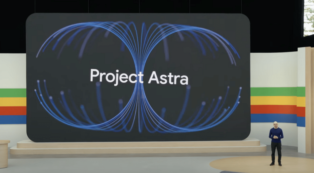 Une capture d'écran d'une présentation de Google présentant le "Project Astra". Un orateur se tient sur une scène devant un grand écran affichant le texte "Project Astra" entouré de motifs graphiques bleu néon en forme de boucle. L'arrière-plan de la scène comporte des bandes horizontales de couleurs vert, jaune, rouge et bleu, caractéristiques de l'identité visuelle de Google.