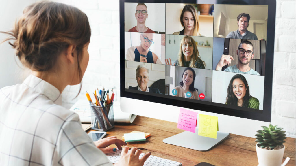 Une femme en télétravail participant à une réunion en visioconférence avec plusieurs collègues affichés sur l'écran de son ordinateur. Elle est assise à un bureau avec des fournitures de bureau et une plante en pot à côté de l'écran.